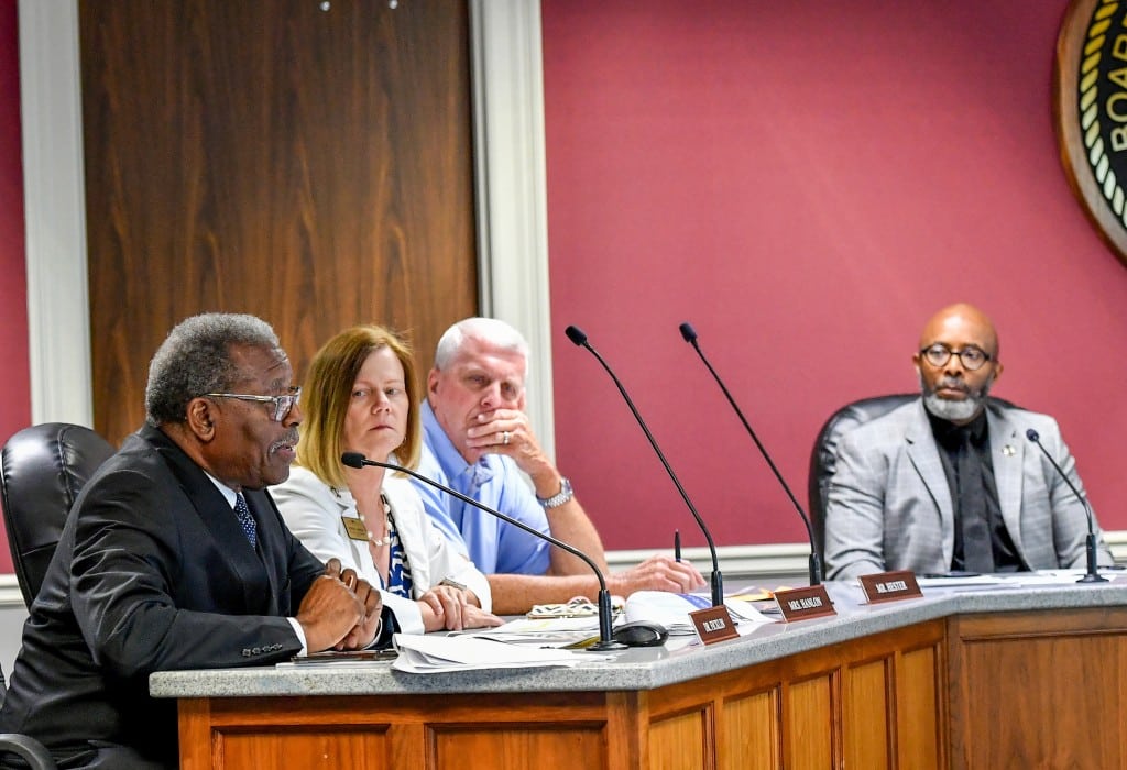 A man speaks into a microphone at his wooden desk while three other people look on.