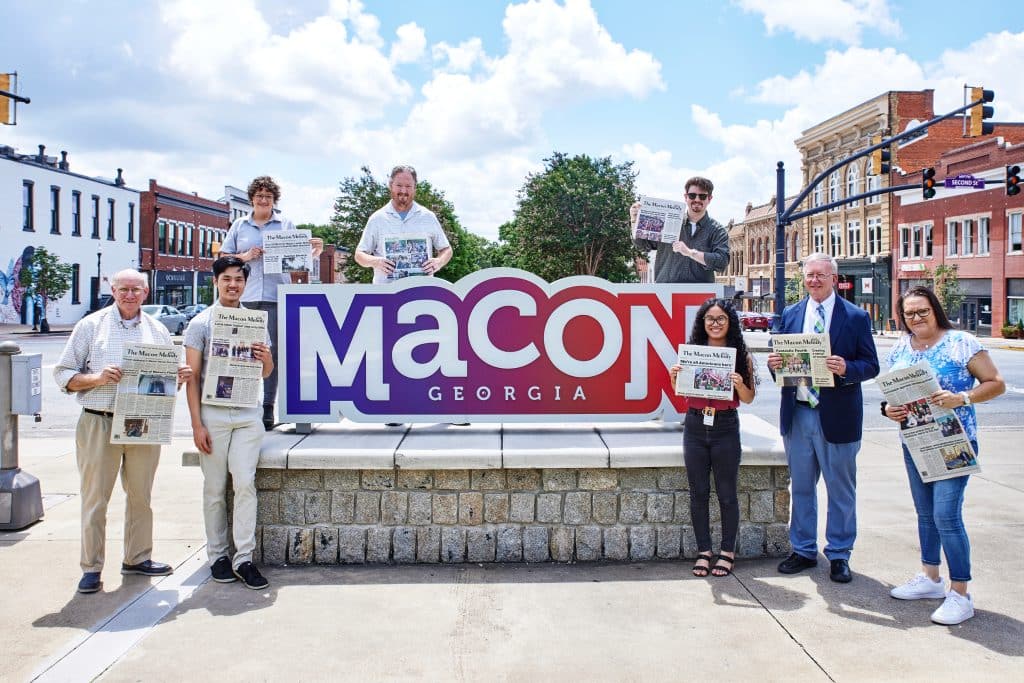 The image shows a group of nine people standing and posing with copies of a newspaper called “The Macon Melody” in front of a large sign that says “Macon Georgia” in bold purple, white, and red letters. The sign is set in a historic downtown area with brick buildings and a clear blue sky with scattered clouds in the background. Some individuals are standing on the ground while others are slightly elevated behind the sign. The mood is cheerful and community-focused.